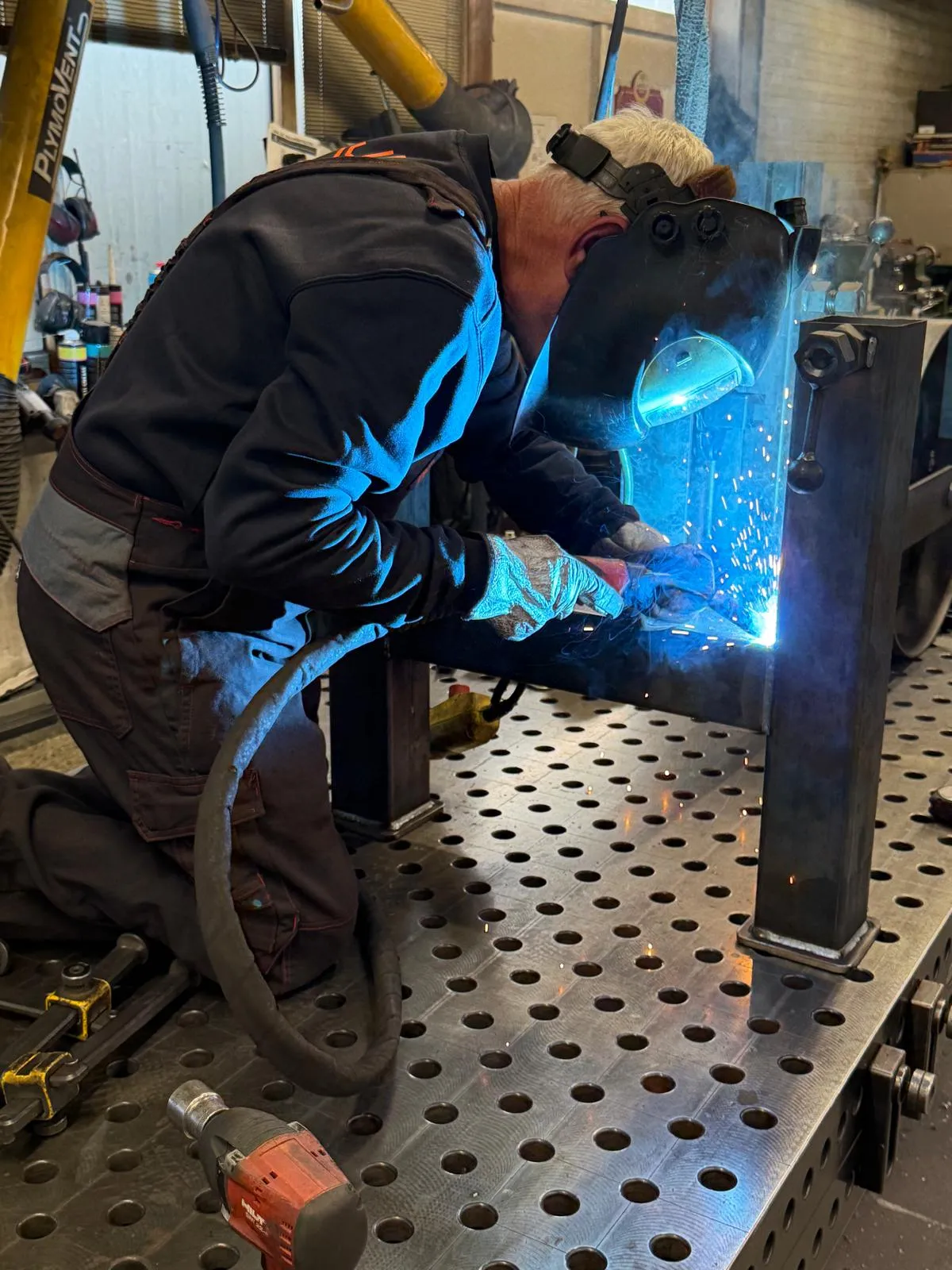 Welder joining a steel table base on a perforated welding table in the workshop