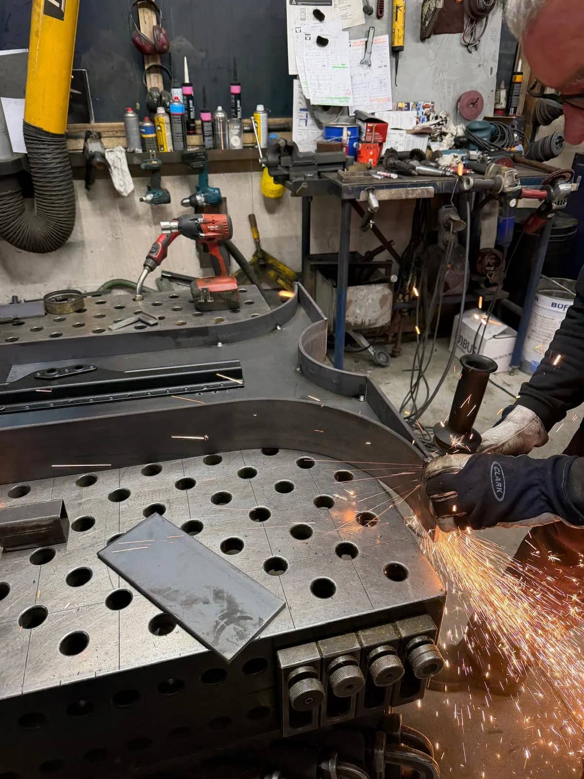Curved steel table base being ground with a shower of sparks on a perforated welding table