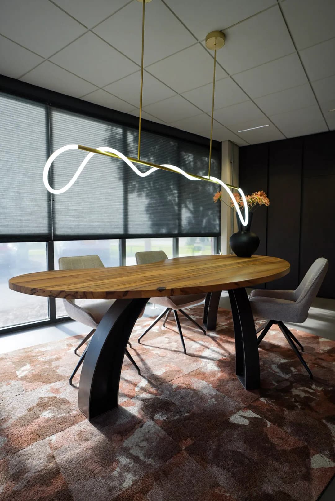 Oval solid wood table on two black arched steel legs with grey chairs under a curved light in an office interior